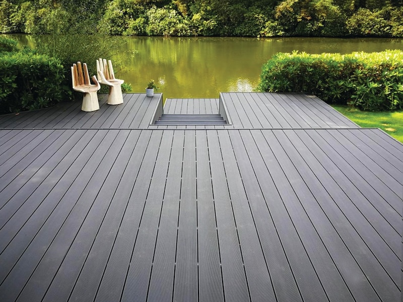 Large grey composite decking area by a lake with unique wooden chairs and greenery.