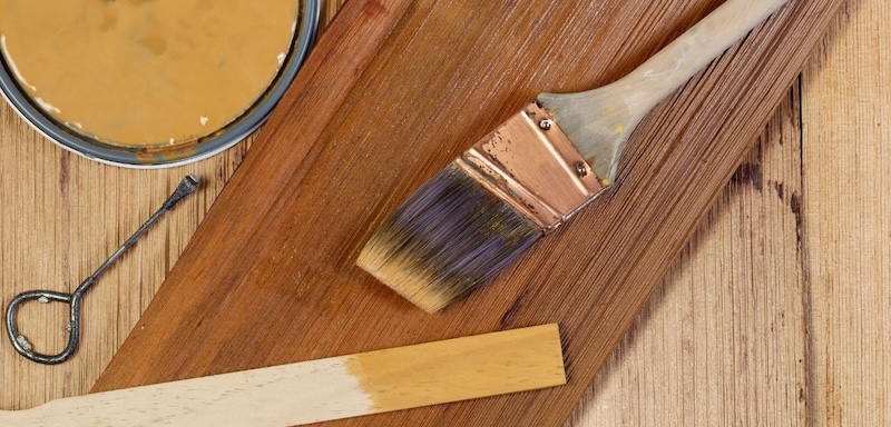 Paintbrush and wood stain on timber board showing staining process for wooden decking