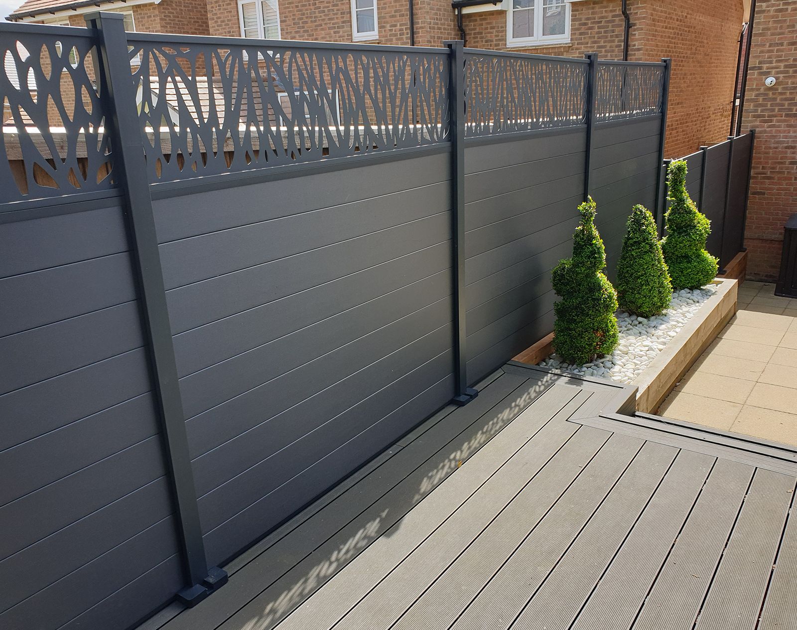 Dark grey composite fencing with decorative slatted top panels, installed alongside raised decking and low-maintenance planting with white stones and spiral topiary.