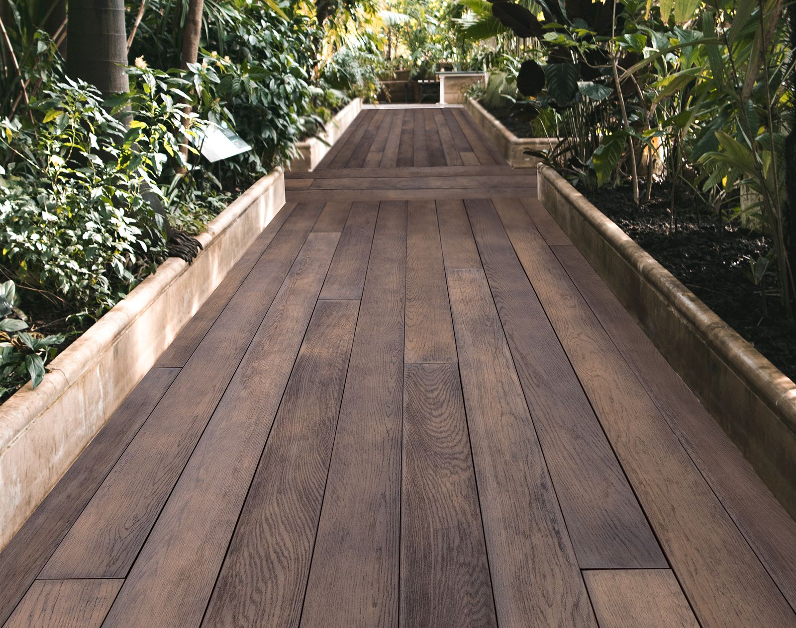 Millboard decking in a rich wood-effect finish used as a walkway through a tropical indoor garden with raised planting beds.