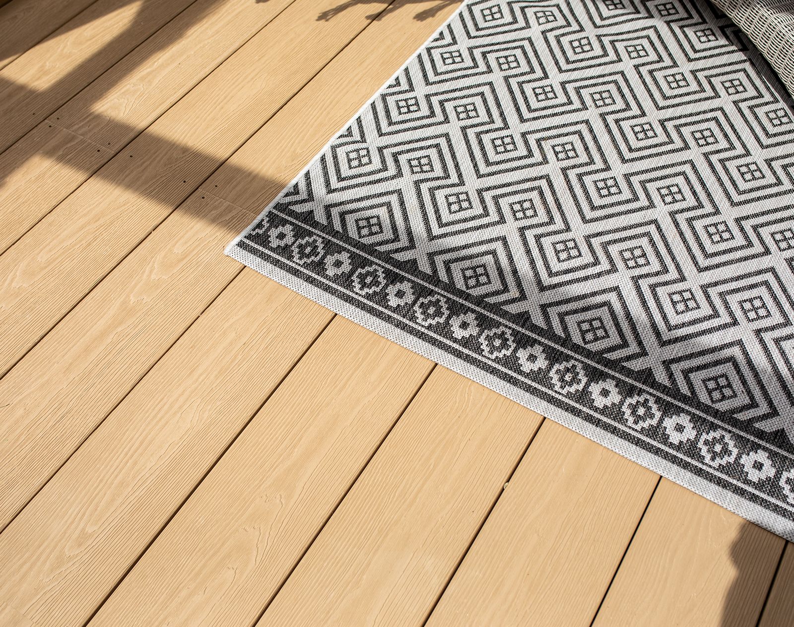 Close-up of light oak-effect Nordeck PVC decking boards with a patterned outdoor rug partially covering the surface in bright sunlight.