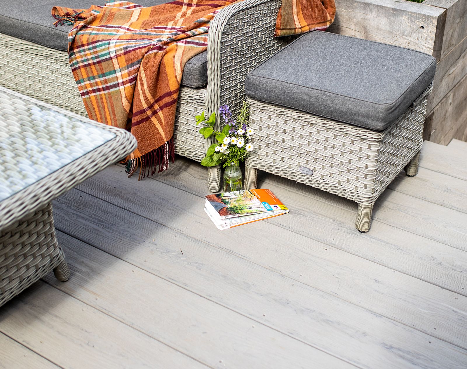 Stylish garden seating area with wicker furniture and light grey Nordeck PVC decking, featuring a blanket, flowers, and magazines for a cosy outdoor vibe.