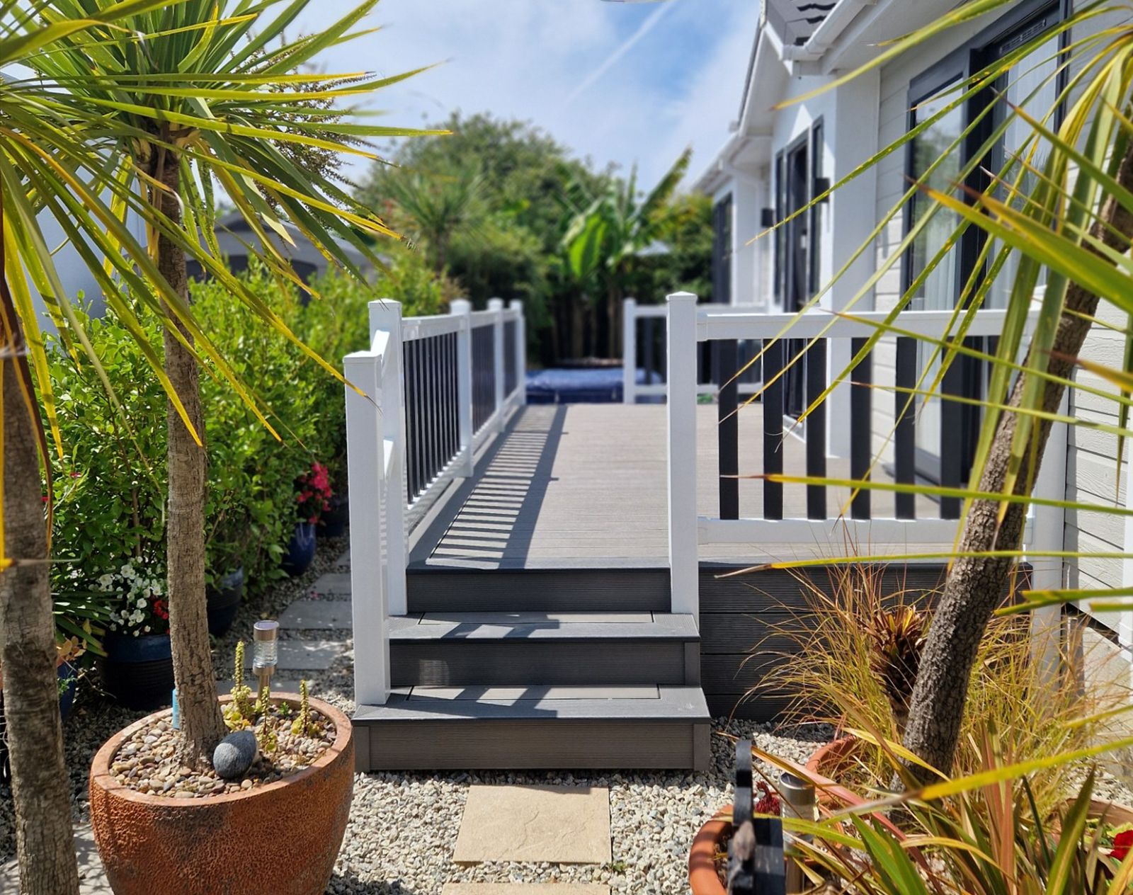Raised composite decking with white and black balustrade in a modern garden with palm trees, gravel landscaping, and a coastal-style home exterior.