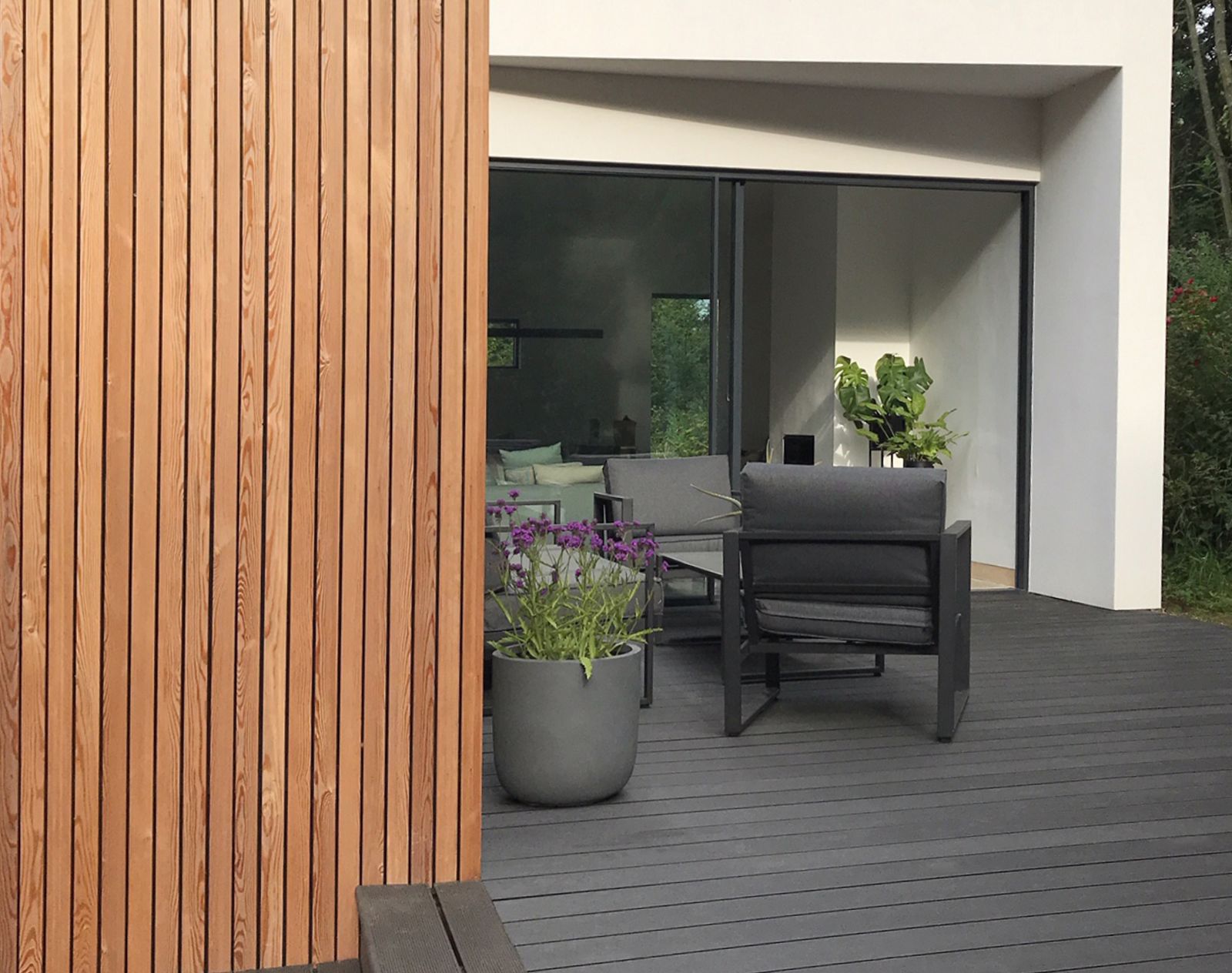 Dark grey composite decking outside a modern home with vertical timber cladding, minimalist garden furniture, and large sliding glass doors.