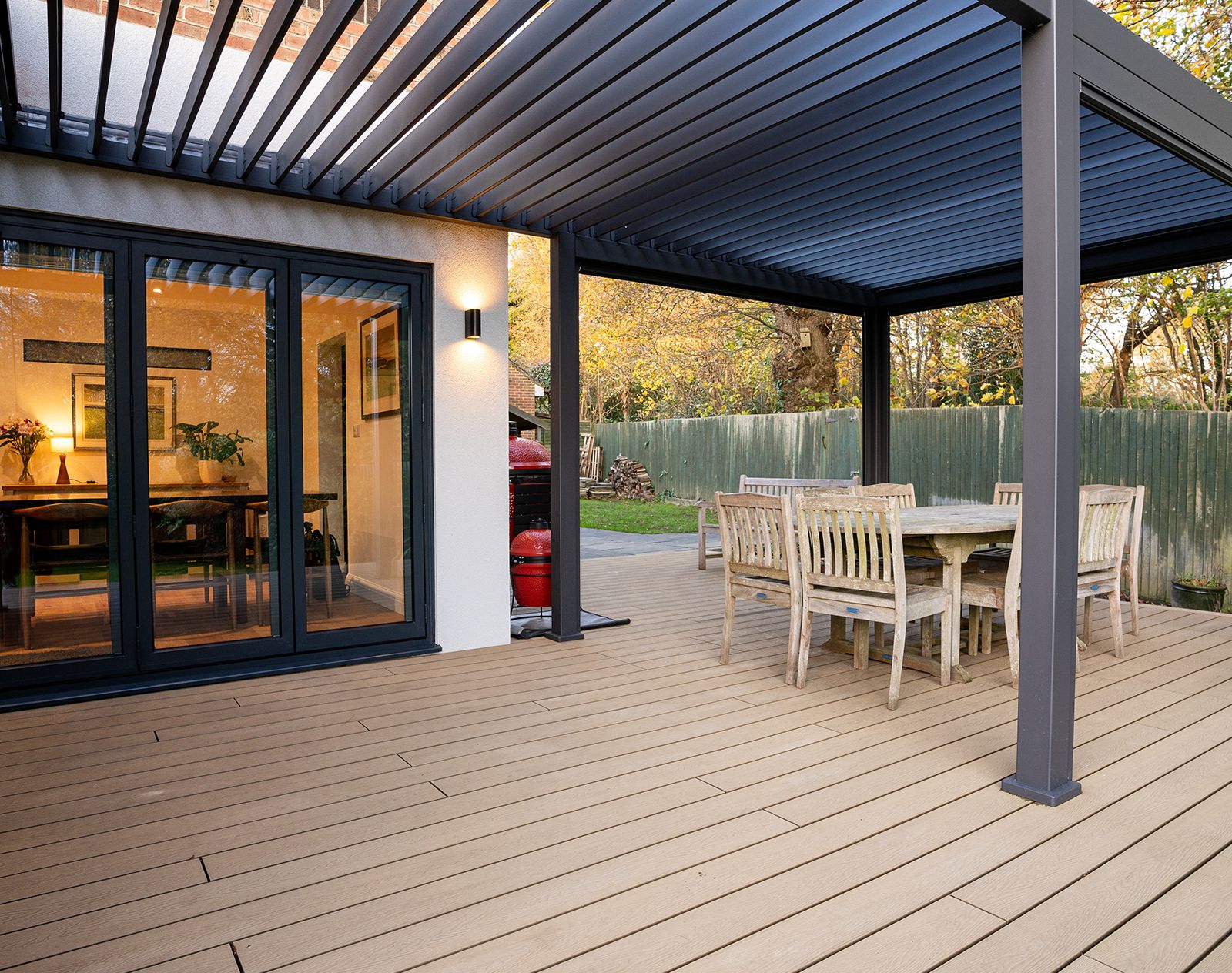 Light oak-effect composite decking under a modern aluminium pergola, with rustic wooden dining furniture and sliding patio doors leading to a garden.