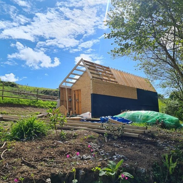 The exposed building during construction reveals large wooden beams before steel corrugated sheets are applied.