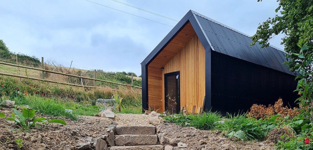 A large garden room with corrugated steel cladding in black and timber slats. Surrounded by fields with stone steps