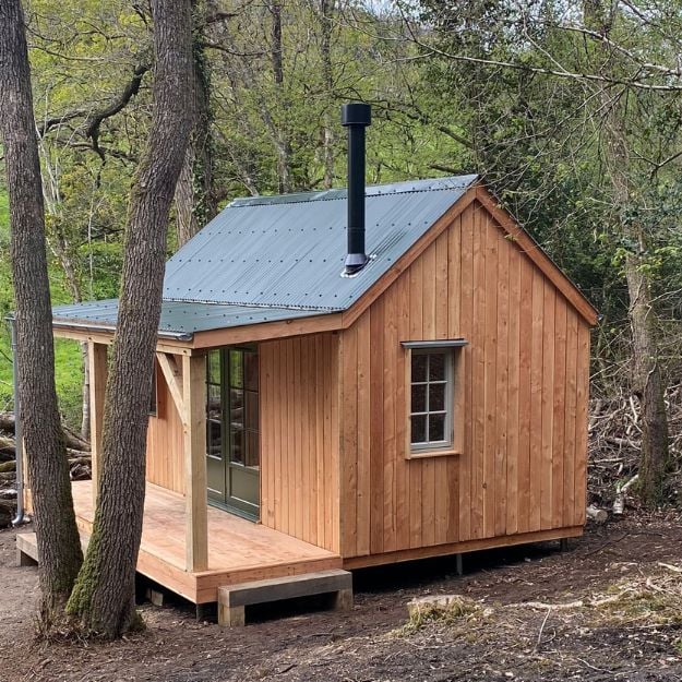 A small wooden cabin in the woods, with an exterior chimney flute and a Juniper Green Corrugated Roof.